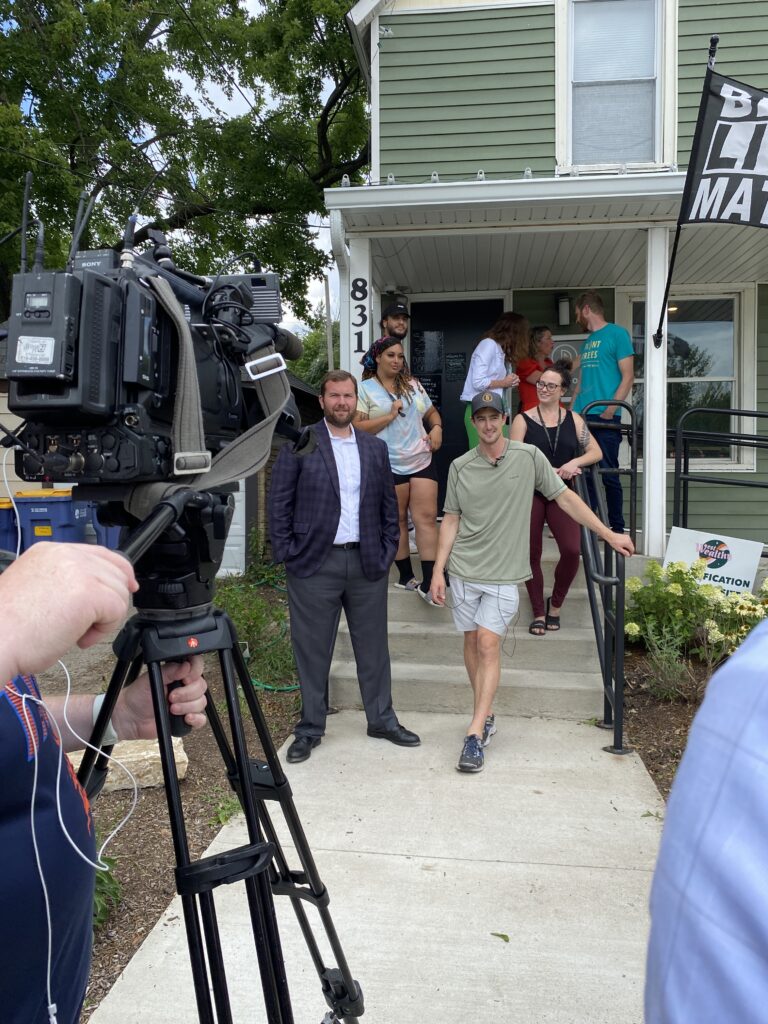 Two men stand in from of a green house with other people in the background. The building is Pharmhouse Wellness. There is a TV camera in the foreground of the photo.